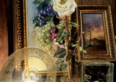 A white rose in a glass vase amidst vintage paintings and a decorative brass plate.