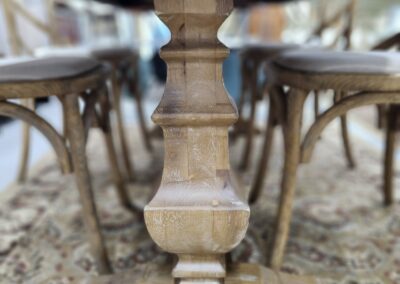 Close-up of a wooden table leg on a patterned rug with chairs in the background.