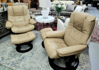 Two beige cushioned swivel chairs with matching ottomans in a furniture store.
