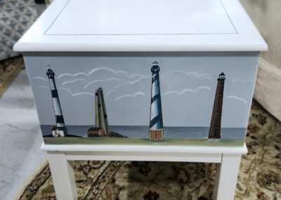 Wooden chest with lighthouse paintings on the front, standing on white legs.