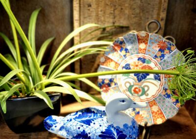 A blue and white ceramic peacock figurine beside a colorful decorative fan and a plant.
