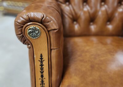 Close-up of a brown leather armchair's armrest with decorative button and stitching.
