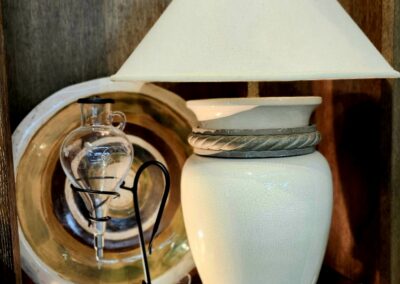 White ceramic lamp with a beige lampshade and decorative objects behind.
