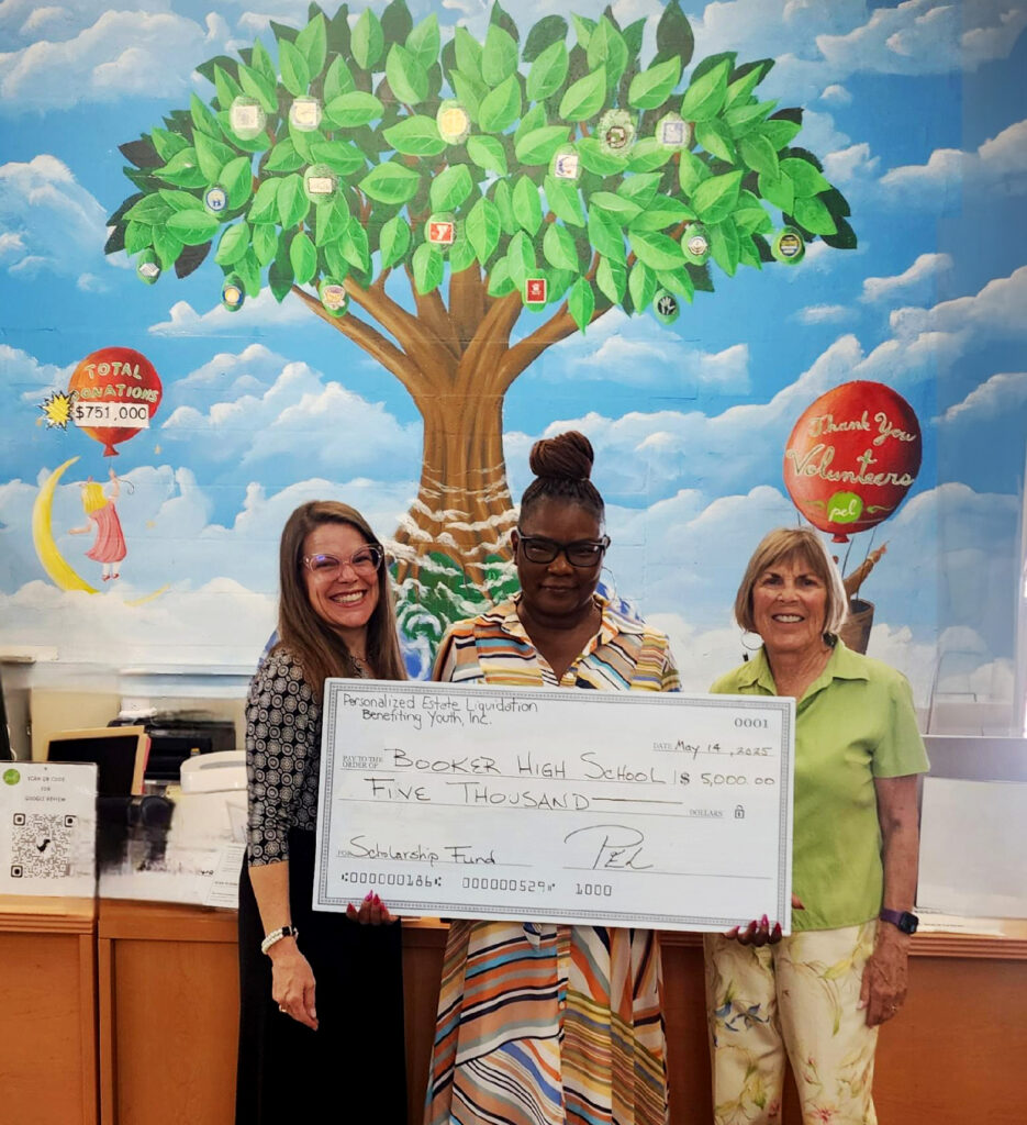 Three women holding a large check in front of a colorful mural with hot air balloons.