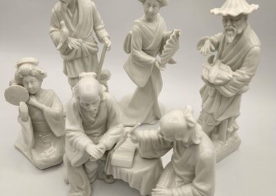 A collection of seven white porcelain figurines depicting traditional Asian attire and activities.