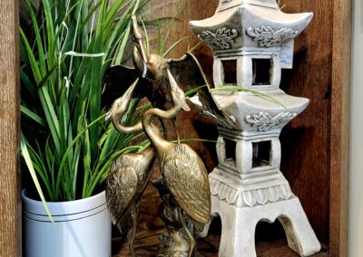 Decorative bird statue with plant and stone lantern on wooden shelf.
